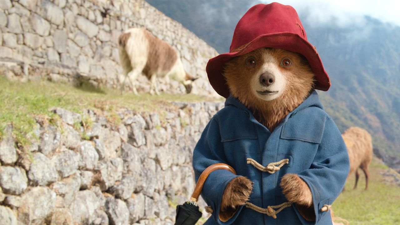 Paddington the bear wearing a blue jacket and red hat next to some Himalayan goats in Paddington in Peru