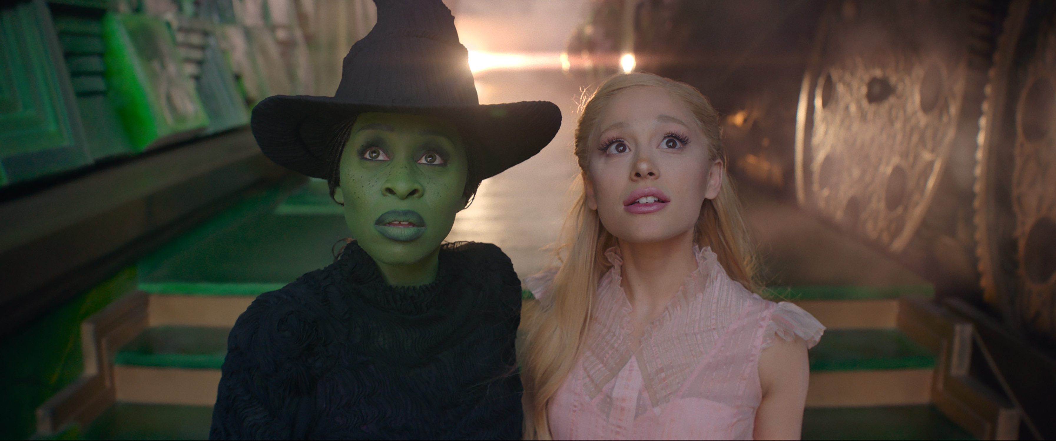 Elphaba, a green-skinned woman, and Glinda, a blonde woman in pink, look wide-eyed at something in Wicked