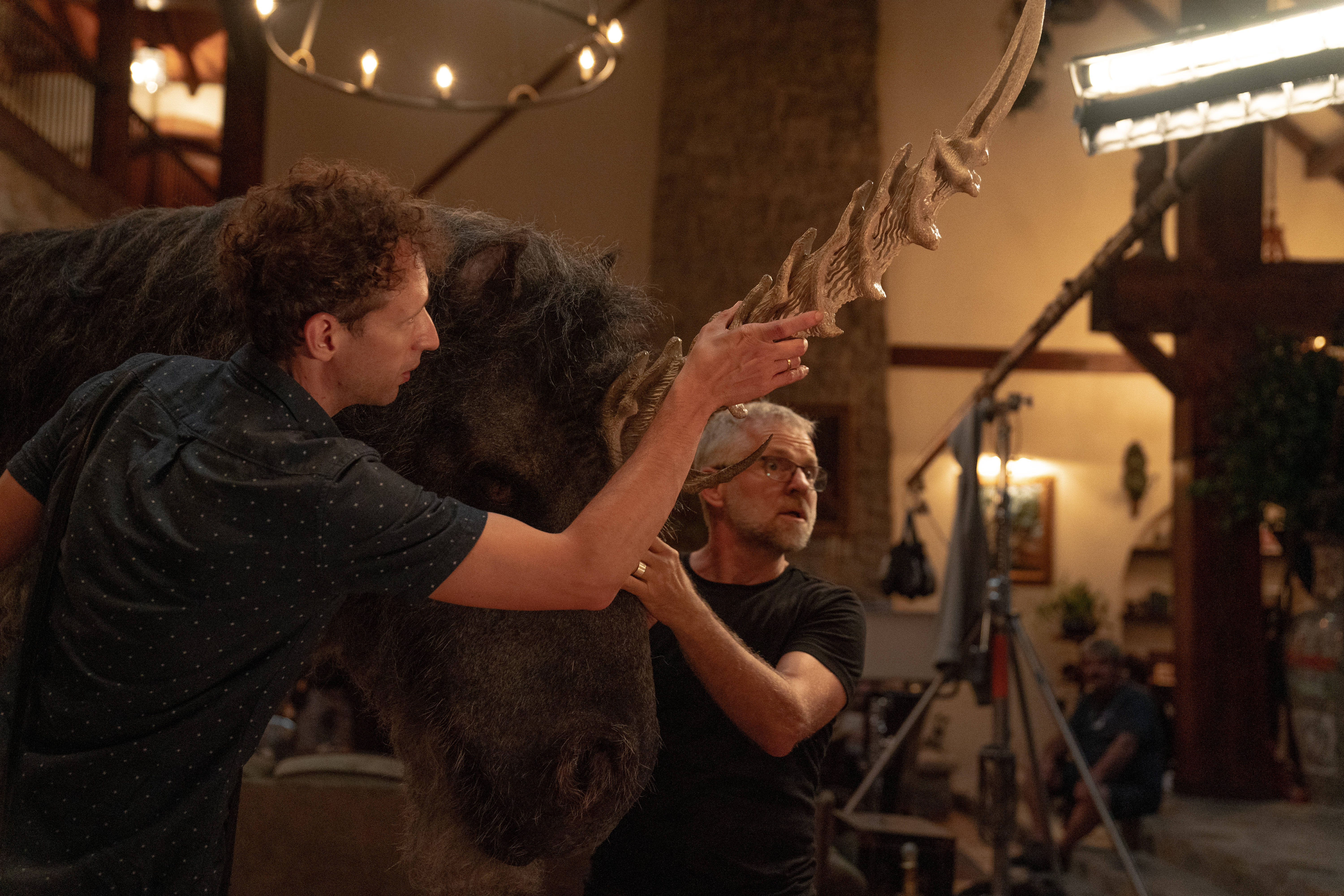 Alex Scharfman and crew adjust a prop unicorn head on the set of Death of a Unicorn. They’re inside a spacious mansion, with film equipment visible around them. The unicorn’s fur is purple-black, and its enormous horn is fearsomely ridged, with sharp, curving smaller spikes at its base.