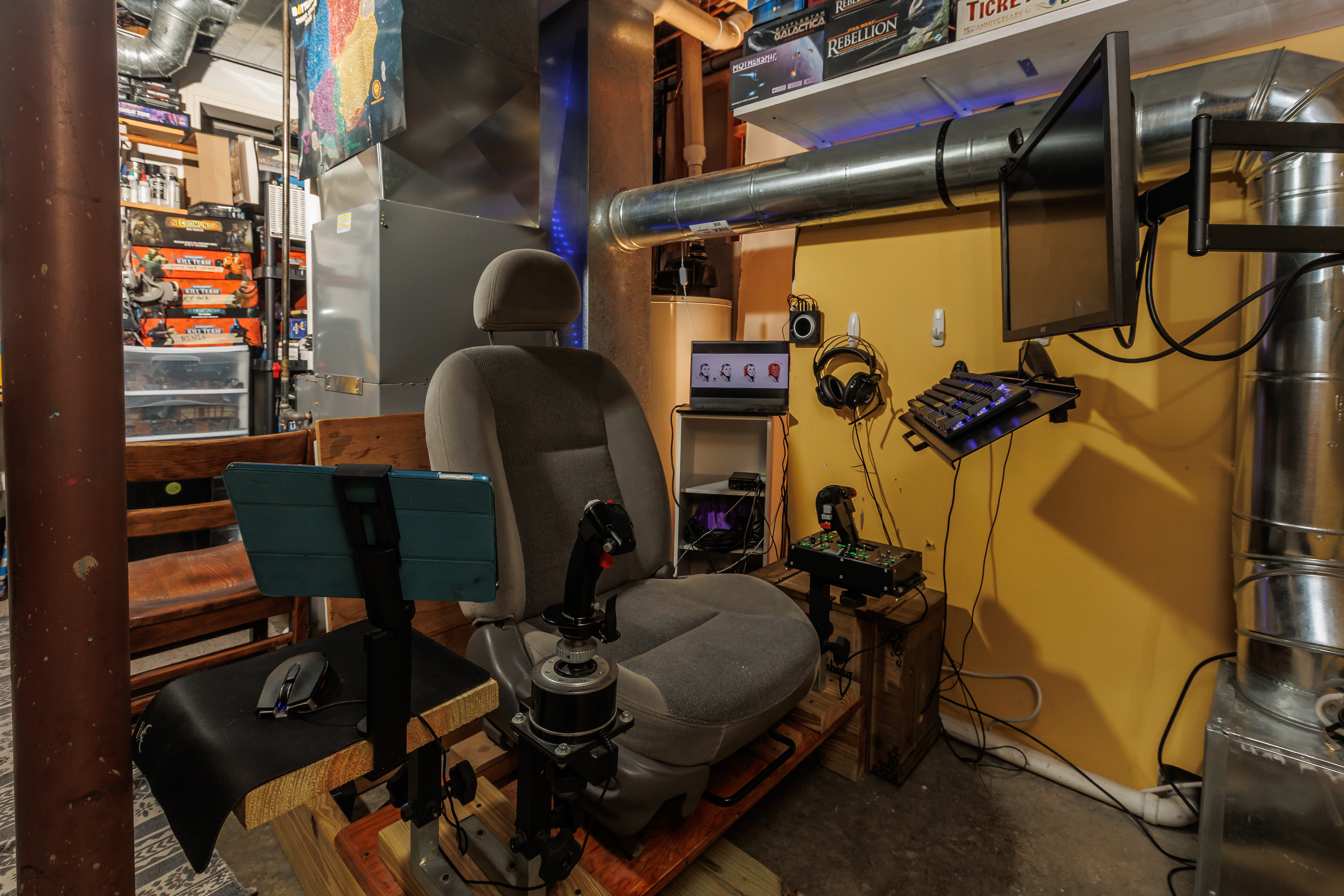 A bucket seat seat wedged in between a furnace and a water heater, below a shelf packed with board games.