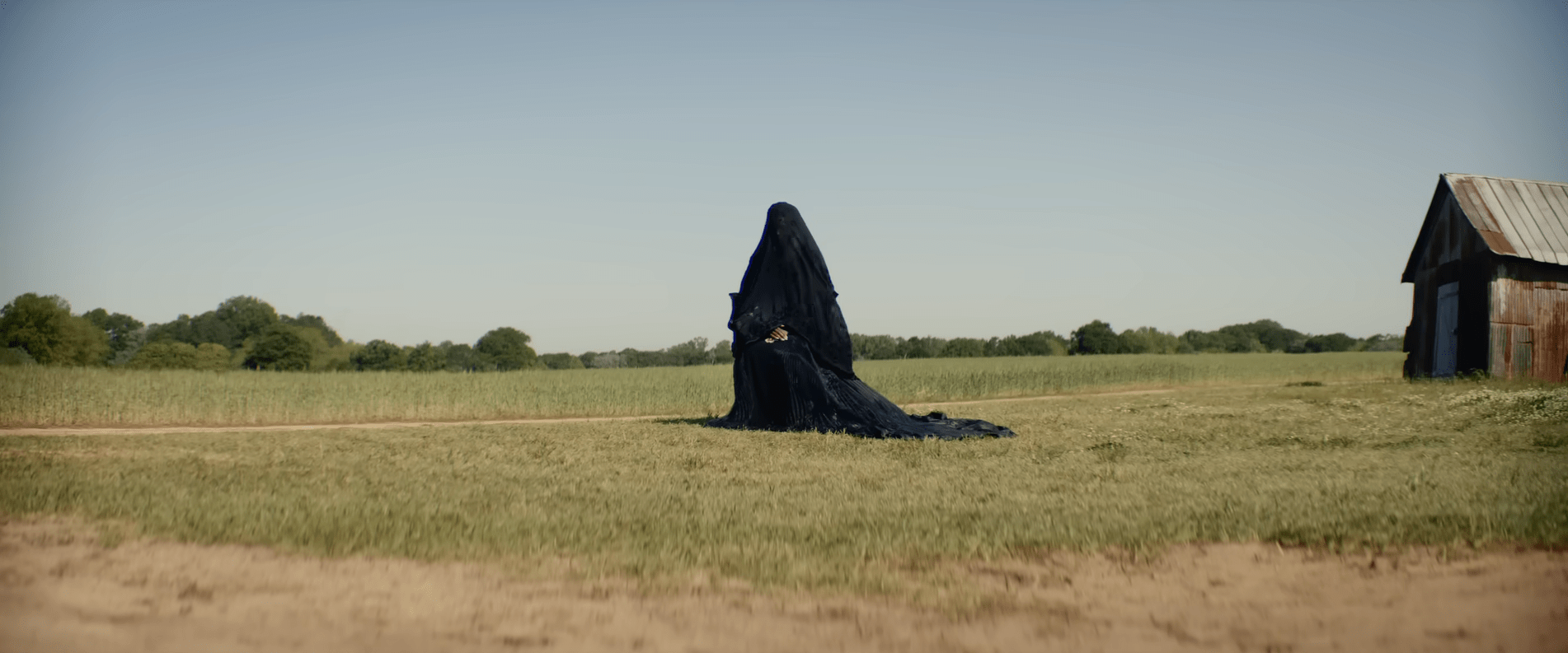 A woman in a black shroud sits alone in an open yard with a barn nearby in The Woman in the Yard