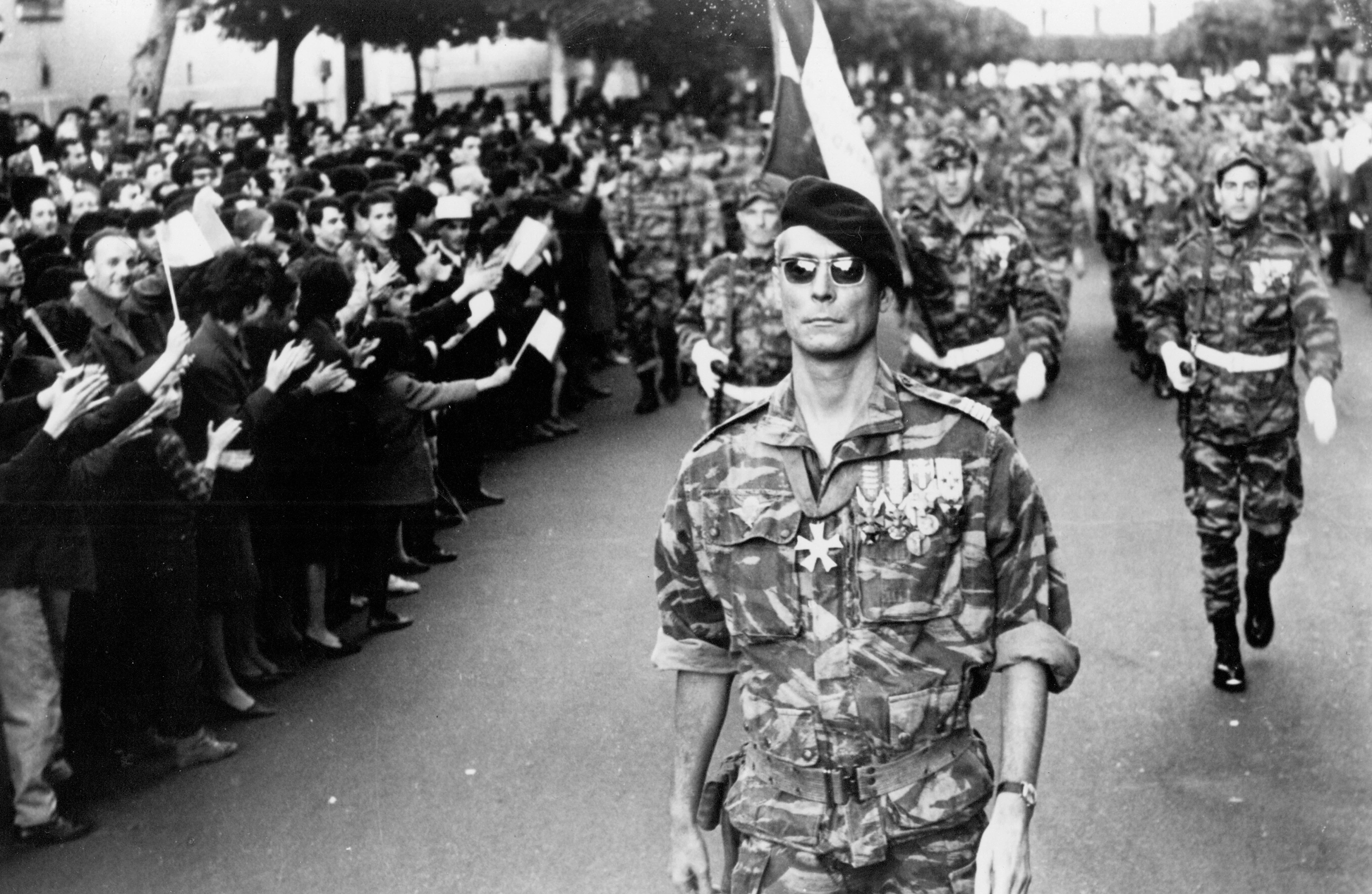 A soldier wearing sunglasses and a beret marching in uniform down a street flanked by people waving flags and cheering in The Battle of Algiers.