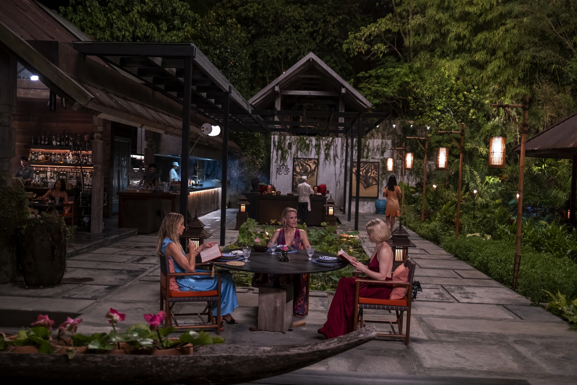 Three White Lotus women sitting at a table talking to each other and reading books