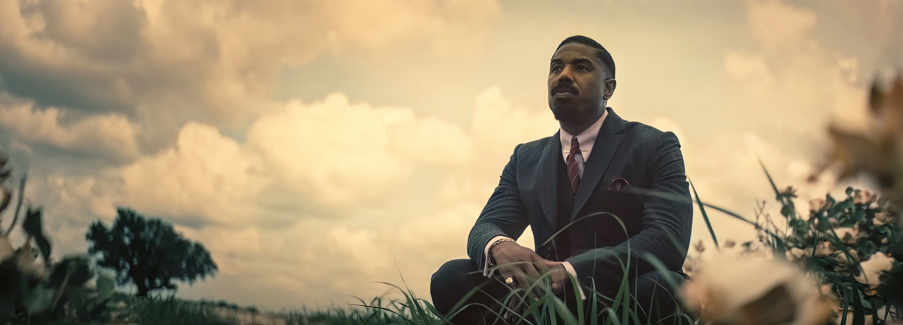 One of the Smokestack twins (Michael B. Jordan) sits in a fine suit in a grassy field and looks up at a cloudy sky in Ryan Coogler’s Sinners