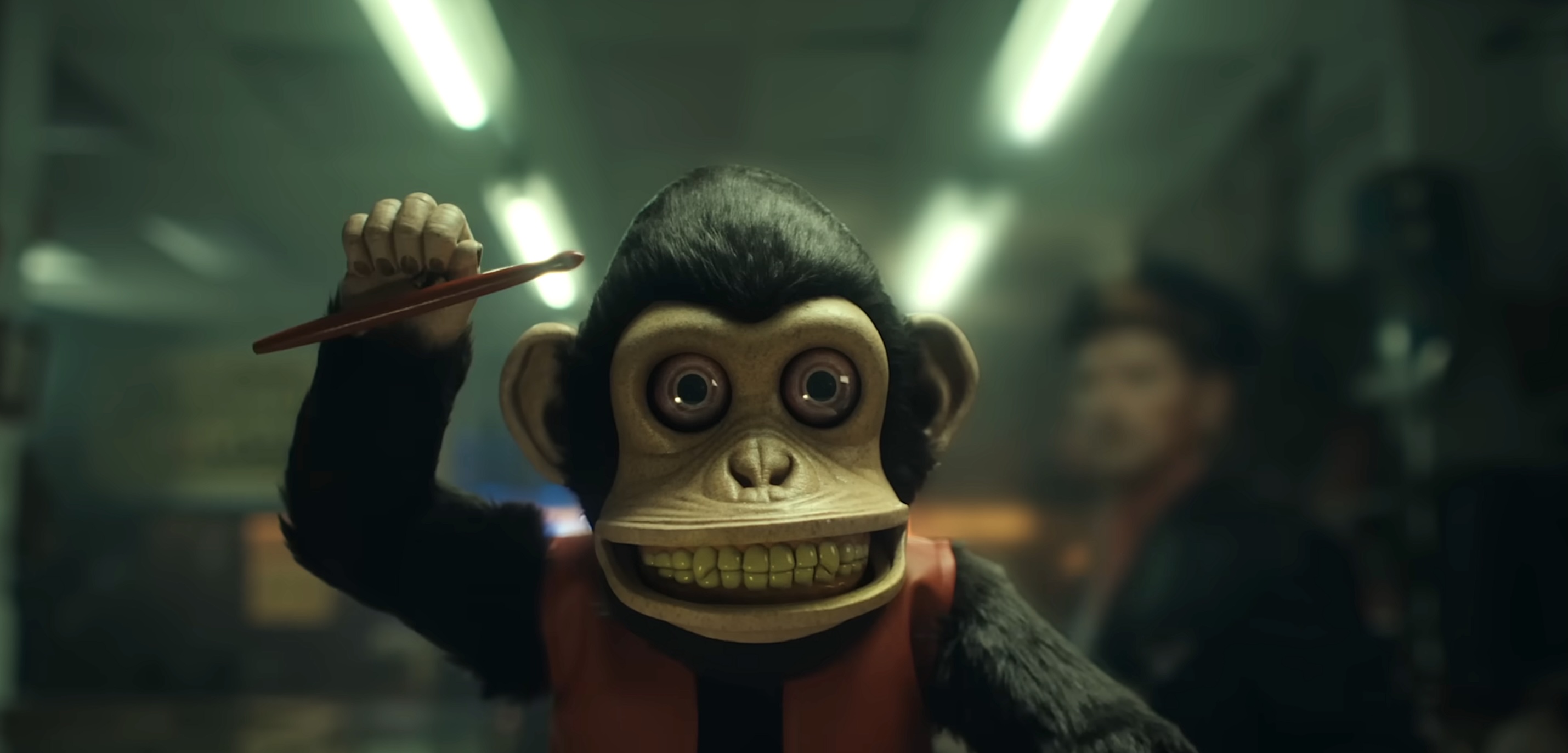 A grinning toy wind-up monkey with one drumstick raised in the air, from the trailer for Osgood Perkins’ The Monkey