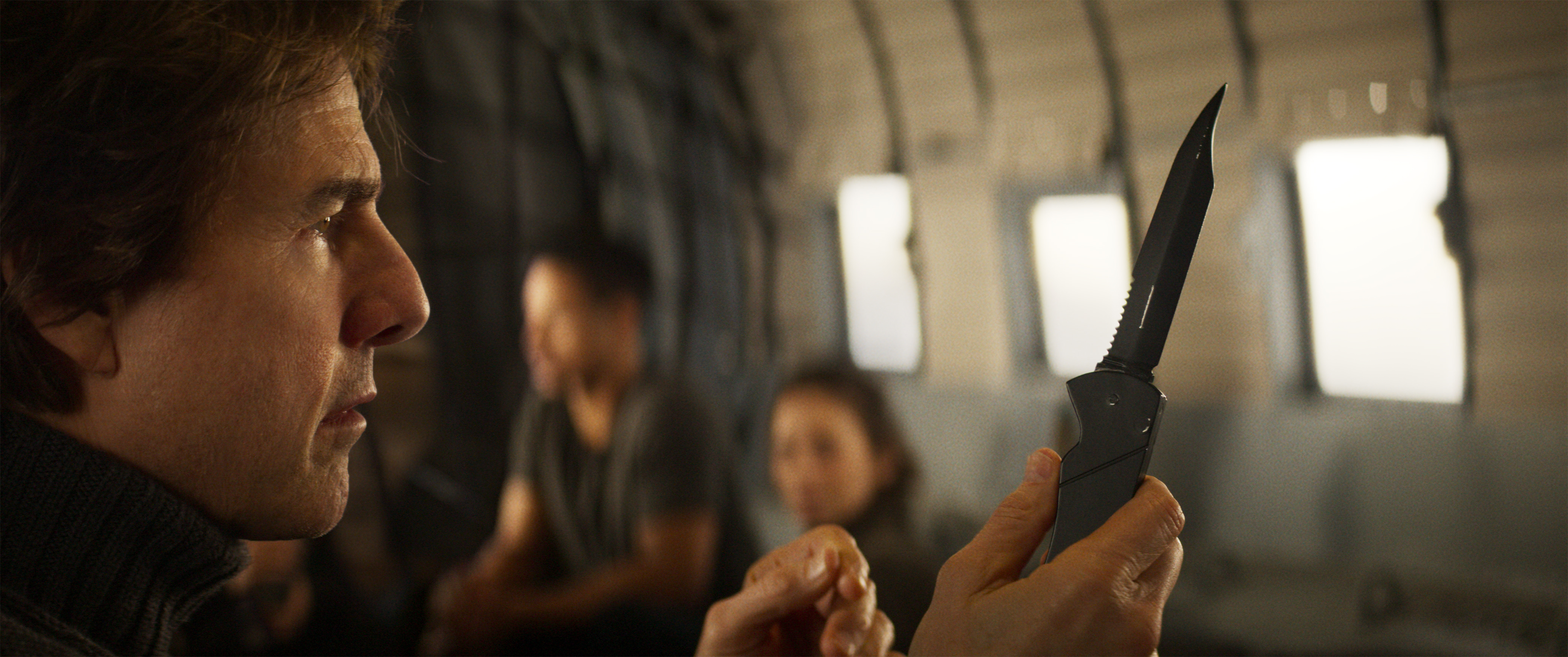 In a scene from Mission: Impossible — The Final Reckoning, Ethan Hunt (Tom Cruise), sitting in a cargo plane with other characters out of focus in the background, holds up the tactical knife he dropped during the big Langley heist in 1996’s Mission: Impossible