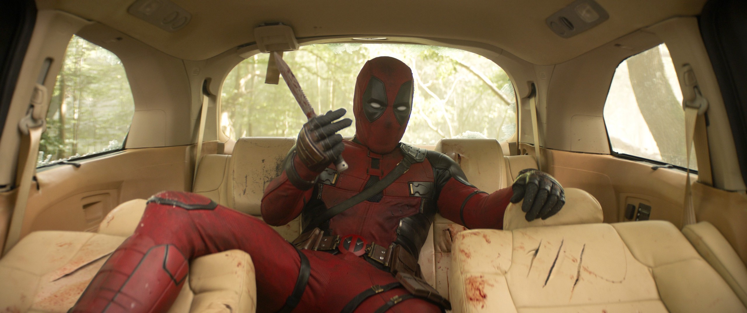Deadpool (Ryan Reynolds) sprawls out, legs spread disconcertingly wide, in the back seat of a shredded, bloodied car in Deadpool & Wolverine